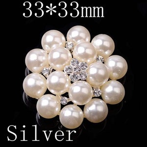 May include: A silver flower-shaped brooch with a center of clear rhinestones surrounded by white faux pearls. The brooch measures 33mm by 33mm.