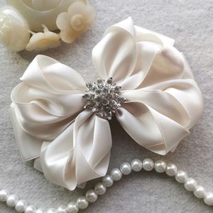 May include: Cream satin bow with a diamante centre. The bow is layered and has a classic design. A strand of white pearls is in the foreground. The bow is a decorative accessory.