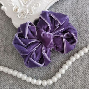 May include: A large purple velvet bow with a satin edge. The bow is lying on a gray fabric surface with a string of white pearls.
