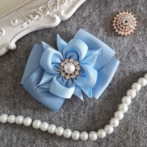 Pode incluir: Uma flor de tecido azul claro com um centro de pérola e strass. A flor está sobre um fundo cinza com uma corda de pérolas brancas.