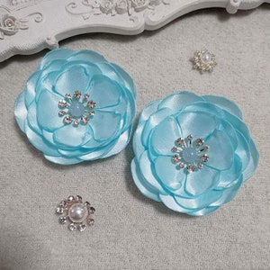 Puede incluir: Dos pinzas para el pelo de satén azul claro con un strass y un centro azul claro. Las flores están superpuestas y tienen un efecto 3D.