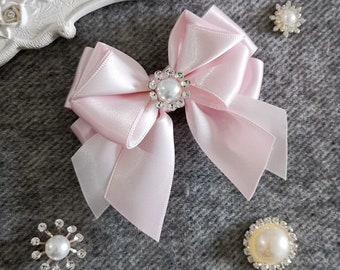Könnte beinhalten: Eine rosa Schleife aus Satin mit einer Perlen- und Strassverzierung. Die Schleife befindet sich auf einer grauen, strukturierten Oberfläche.