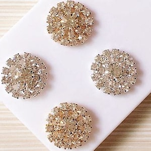 May include: Four gold and clear rhinestone embellishments. Each embellishment is a round, flat, circular design with a central rhinestone surrounded by smaller rhinestones.