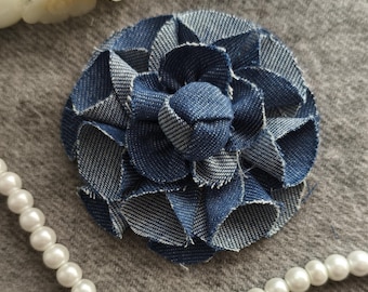 Pode incluir: Broche de flor de ganga com um design de pétalas em camadas. O broche é feito de tecido de ganga azul e branco, criando uma aparência floral texturizada. Um fio de pérolas brancas e flores brancas estão no fundo.