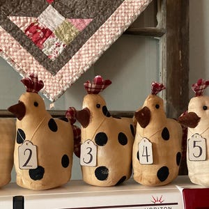 May include: Five fabric chicken figurines in tan and cream, each featuring a brown beak, a red and white checkered comb, and a small tag numbered one through five. Two chickens have black polka dots, while three have a subtle tan checkered pattern.