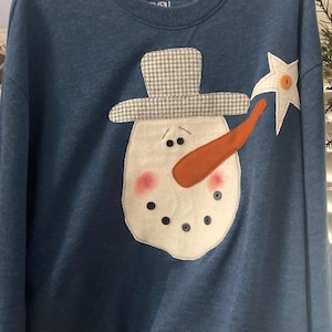 May include: A blue long-sleeve sweatshirt featuring a large snowman applique on the front. The snowman has a grey houndstooth hat, an orange carrot nose, pink cheeks, and dark button eyes and mouth. A white star with an orange button is attached.