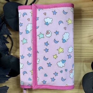May include: A rectangular fabric planner cover in light pink featuring a repeating pattern of yellow stars, blue crescent moons, and white clouds with blue hearts. The cover is edged with a solid dark pink fabric trim and includes a ribbon bookmark.