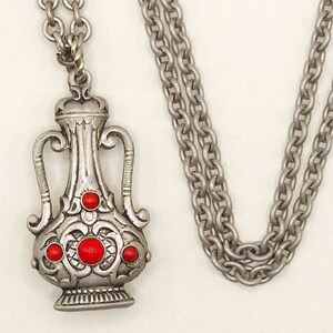 Puede incluir: Collar de plata con un colgante detallado y adornado en forma de jarrón con acentos de piedras preciosas rojas.