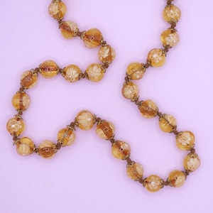 Op de afbeelding: Een lange ketting gemaakt van amberkleurige glazen kralen met ingewikkelde bloemmotieven. De kralen zijn aan elkaar geregen met goudkleurig draad.