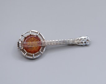 Op de afbeelding: Een zilveren banjo broche met een amberen steen in het midden. De banjo heeft een gedetailleerd ontwerp en is een vintage sieraad.