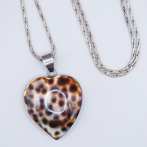 Può includere: Un ciondolo a forma di cuore con una superficie liscia e lucida caratterizzata da un motivo a macchie marroni e crema simile a quello di un leopardo. Il ciondolo è fissato a una catena in metallo argentato con maglie intrecciate.