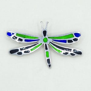 Pode incluir: Um broche de libélula de prata com detalhes em esmalte preto, verde e azul. As asas da libélula estão abertas e o corpo é detalhado com esmalte preto e verde.