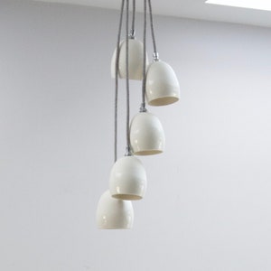 May include: A white ceramic pendant light fixture with six bulbs hanging from the ceiling. The light fixture is in a modern style and is hanging over a wooden table with a bottle of wine and a bouquet of white roses.