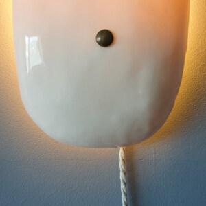 May include: A white ceramic wall sconce with a round shape and a single light bulb. The sconce has a small, round, brass-colored button on the front and a white cord hanging from the bottom.