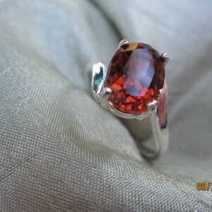 May include: A silver ring with a large, oval-shaped, reddish-brown gemstone. The ring is set on a soft, gray fabric.