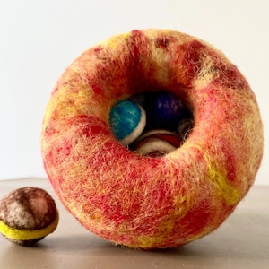 May include: A felted wool donut-shaped container in shades of red, yellow, and orange, filled with small, colorful felt balls. A smaller felt ball with brown and yellow accents sits beside it. The handmade item is on a neutral surface.