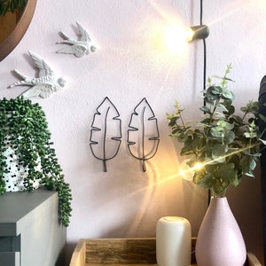 May include: Two black metal leaf wall decorations on a pink wall with a white ceramic bird wall decoration, a potted plant, and a white diffuser on a wooden table.