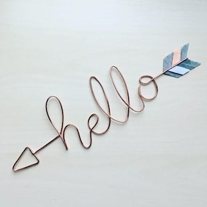 May include: A copper wire sculpture of the word "hello" with an arrow made of felt in shades of grey, pink, and white.