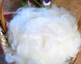 May include: A round wooden basket with a curved handle containing a large, fluffy mound of white raw fiber. Tucked behind the white fiber are several small, artificial decorative flowers in shades of red, purple, and green with brown stems.
