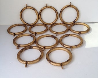 May include: A set of 12 gold-toned metal curtain rings. The rings are round and have a hook on the top. They are arranged in a triangular pattern on a white surface.