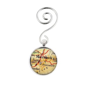 May include: Silver ornament featuring a round map of Marrakech, Morocco. The map is printed on a cream-colored background with red roads and black text. The text on the map reads "Marrakech A.D. 1333" and "Toukbal 13,671".