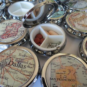 May include: A set of silver pill boxes with vintage map designs. Each box has three compartments and a hinged lid. The maps feature locations like Dallas, San Francisco, and the Isle of Man.