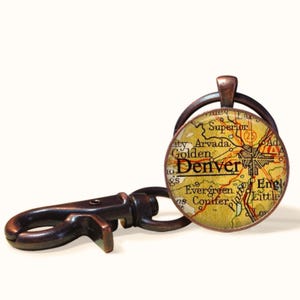 May include: A round keychain featuring a map of Denver, Colorado, with the city name prominently displayed. The map has a vintage, aged appearance with a yellow and brown color scheme. The keychain has a bronze-colored metal finish and a clasp.