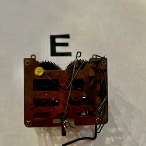 May include: A brass clock movement mechanism featuring exposed gears, a rectangular frame, and various metal levers and wires. The letter E is positioned above the mechanism. The internal components are visible, showing a complex arrangement of wheels and springs.