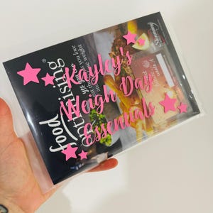 Può includere: Una custodia di plastica trasparente contenente una rivista con il testo "Food Optimising" e "Kayley's Weigh Day Essentials" in rosa. Abbellimenti a forma di stella rosa sono sparsi sul davanti. La rivista presenta immagini di cibo.