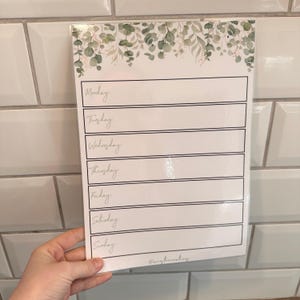 May include: A white weekly planner with a botanical design at the top, featuring green leaves and gold accents. The days of the week are listed in a handwritten font. The planner is held against a white tile background.