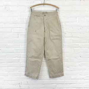 May include: A pair of beige cotton pants with a button fly and a straight leg. The pants have a slight distressed look and are hanging on a wooden hanger.