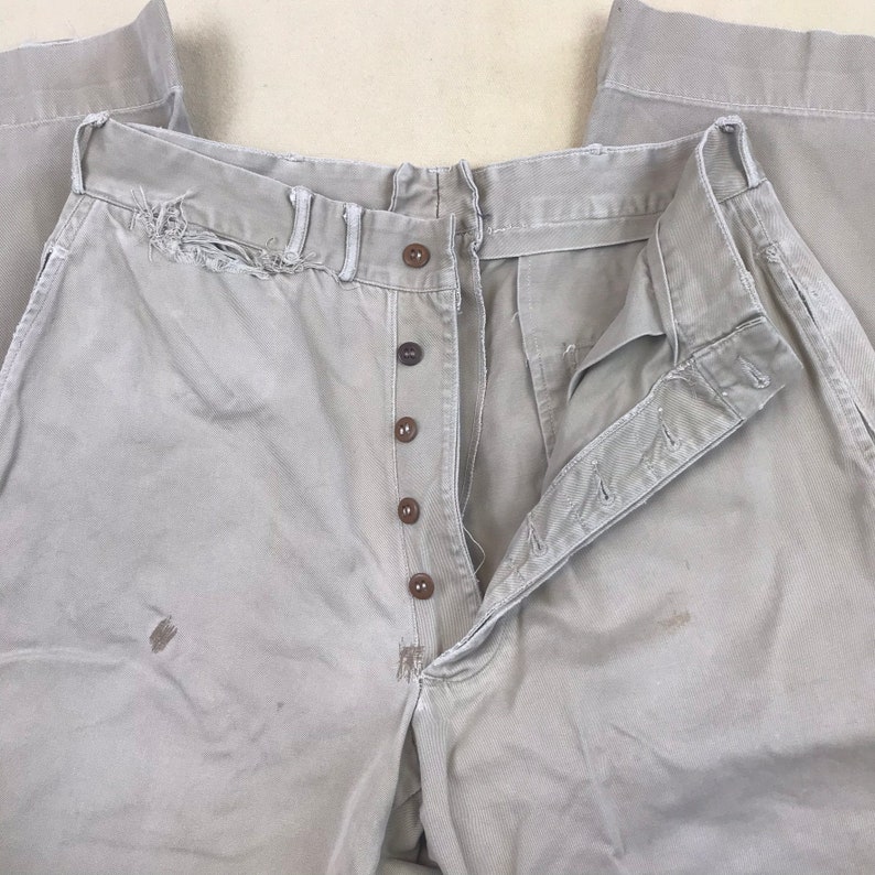 May include: A pair of light brown pants with a button fly and multiple buttons. The pants have a distressed look with some fraying and a few stains.
