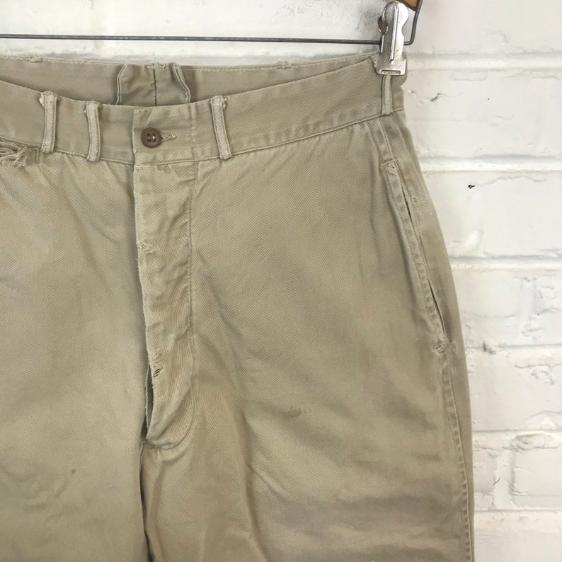 May include: A pair of beige cotton twill pants with a button fly and a single front pocket. The pants have a worn look and are likely vintage.