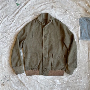 May include: A green wool bomber jacket with a ribbed knit collar, cuffs, and waistband. The jacket has two large flap pockets and a button closure.
