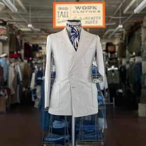 May include: A light beige double-breasted blazer with wide peak lapels, two front patch pockets, and a single chest pocket. The blazer is styled with a dark blue neck scarf featuring a white and yellow leaf pattern. The garment is displayed on a mannequin.