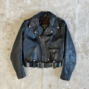 May include: Black leather motorcycle jacket with a classic asymmetrical zip closure, featuring a belted waist, multiple pockets, and silver-tone hardware. The jacket has a vintage aesthetic and a quilted lining.