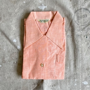 May include: A folded peach-colored button-down shirt with a pointed collar and a single pocket. The shirt has a subtle speckled pattern and a gold-colored button. The label reads "Turner Jogs".