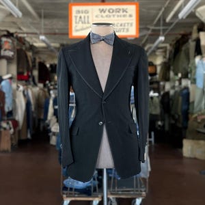 May include: A black blazer with a two-button front closure, wide lapels, and two flap pockets, displayed on a mannequin. The blazer is paired with a small, grey and white patterned bow tie at the neck. The garment is presented in a retail setting.