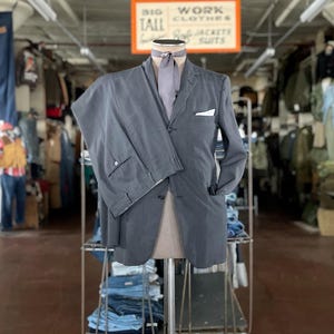 May include: A gray suit displayed on a mannequin, featuring a jacket with a pocket square and matching trousers. The suit is paired with a gray tie. The background includes racks of clothing and a sign that reads "BIG TALL WORK CLOTHES."