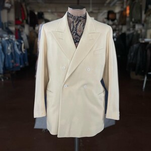 May include: Cream-colored double-breasted blazer with four buttons and a notched lapel. The jacket has a classic design and is displayed on a mannequin. A patterned scarf is visible at the neck. The background shows a clothing store.