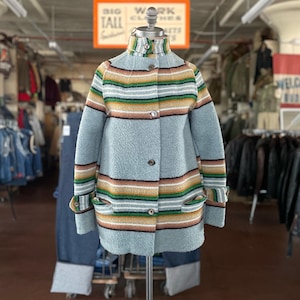 May include: A vintage-style coat in a light blue hue, featuring horizontal stripes in brown, green, yellow, and white. The coat has a high collar, button closures, and pockets. It is displayed on a mannequin in a clothing store.