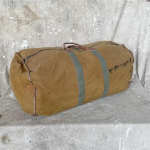 May include: A large, tan canvas duffel bag with two light green stripes and brown leather handles. The bag has a cylindrical shape and appears to be vintage or well-used, with visible wear and tear. The background is a mottled, neutral color.