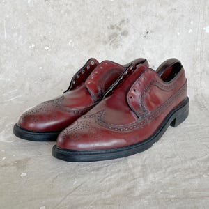 May include: A pair of burgundy leather brogue shoes with black soles. The shoes feature decorative perforations and wingtip detailing. The shoes are displayed on a neutral, textured surface.