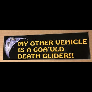 May include: Black rectangular sticker with yellow text that reads "MY OTHER VEHICLE IS A GOA'ULD DEATH GLIDER!!" A blue and silver graphic of a Death Glider is on the left. The sticker is on a brown surface.