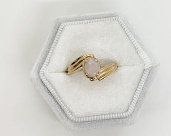 May include: A gold ring featuring an oval, pale gemstone. The ring has a distinctive design with a split band that wraps around the gemstone. The ring is presented in a grey, hexagonal jewellery box.