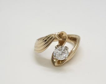 Puede incluir: Un anillo dorado con un diseño en espiral. El anillo presenta una gema grande, transparente y de talla redonda sujeta en un engaste de garras. La banda tiene un patrón texturizado y acanalado. El anillo está sobre un fondo blanco.