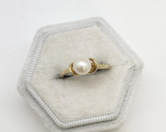 May include: A gold ring with a single, round pearl set in the center. The ring is displayed in a gray, hexagonal jewelry box with a soft, velvety interior. The ring's design features a knot-like detail on either side of the pearl.