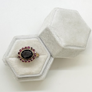 May include: A gold ring with a large black oval gemstone surrounded by a halo of smaller pink gemstones. The ring is displayed in a white velvet ring box.