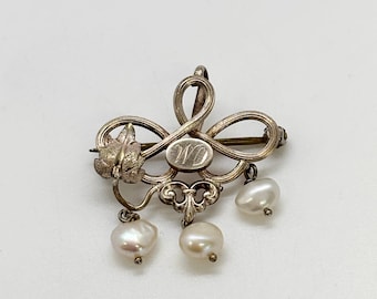 Puede incluir: Broche plateado con diseño floral y de volutas, con tres perlas colgantes de forma irregular. El broche tiene un centro ovalado con las iniciales "WD" grabadas.