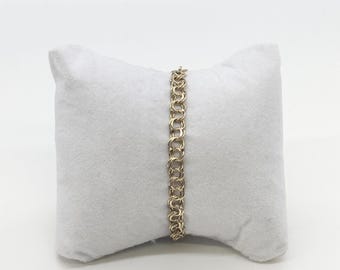 May include: A gold-colored double-link chain bracelet displayed on a white, plush pillow. The bracelet's links are closely connected, creating a textured appearance. The background is a plain white surface, highlighting the jewelry.